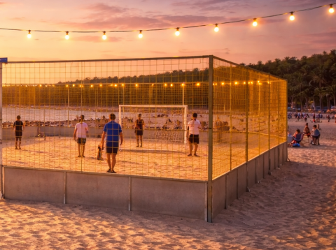 Beach Soccer Court bei Sonnenuntergang am Strand, Spieler auf Sandfeld, umlaufendes goldenes Netz, Lichterkette, Liegestühle im Vordergrund