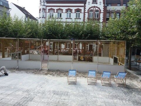 Beach Soccer Court aus Aluminium mit Sandfeld und goldenem Netz auf städtischem Platz, Liegestühle mit Werbung im Vordergrund, Altbaufassade im Hintergrund