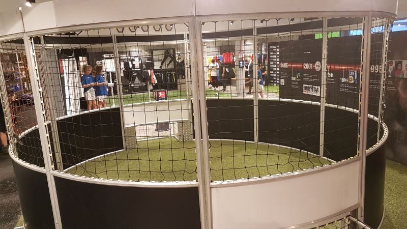 Runder Soccer Cage aus Aluminium in Messehalle, Innenansicht mit Kunstrasenboden und umlaufendem Netz, Besucher im Hintergrund