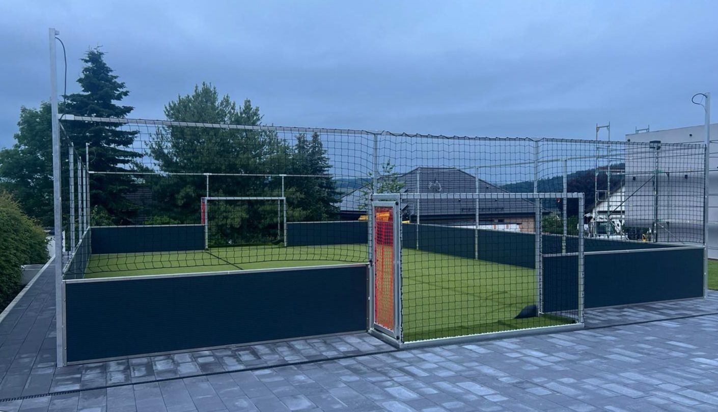 Stationärer Soccer Court Arena Comfort auf gepflastertem Platz in urbaner Umgebung, anthrazitfarbene Banden, umlaufendes Netz, Zugangstür mit Beleuchtung, Abenddämmerung