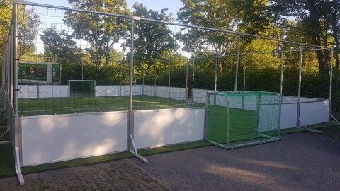 Mobile Soccer Courts Arena Mobil 10×15 m kaufen, direkt vom Hersteller Soccer Eventsport Mobiler Soccer Court Arena Mobil 10×15 m im Freien, weißes Bandensystem, umlaufendes Netz, grüne Baumkulisse, Sonnenschein