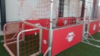 Speedkick mieten mit individuellem Branding, Aluminium, Soccer Eventsport Speedkick-Anlage mit zwei Aluminiumtoren und umlaufendem rotem Bandensystem mit RB-Leipzig-Branding, aufgestellt auf Kunstrasen in einer Halle, Frontalansicht