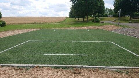 Großes Sportfeld aus verlegtem Kunstrasen mit weißen Markierungslinien im Freien, Ackerlandschaft und Bäume im Hintergrund