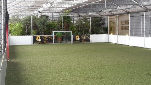 Stationärer Soccer Court mit weißen Banden, umlaufendem Netz und Überdachung, grüner Kunstrasen, Fußballtor mit Vereinsbranding in der Mitte, Innenansicht von der Längsseite