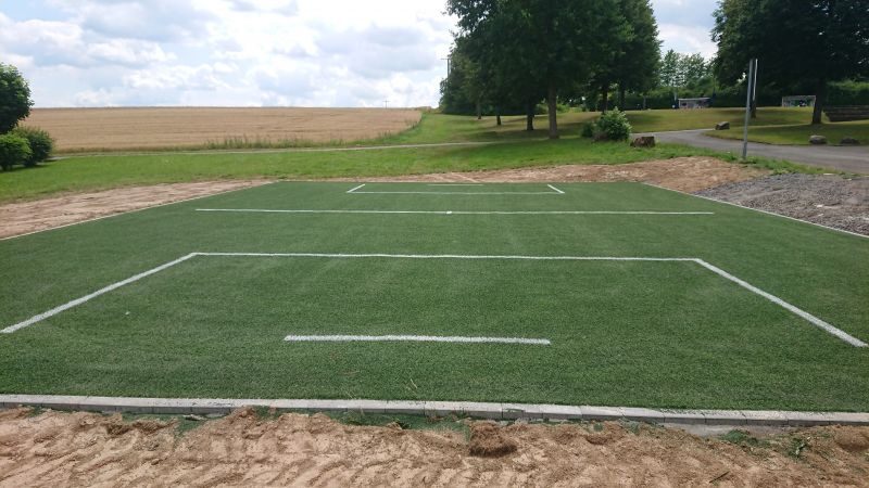 Großes Sportfeld aus verlegtem Kunstrasen mit weißen Markierungslinien im Freien, Ackerlandschaft und Bäume im Hintergrund