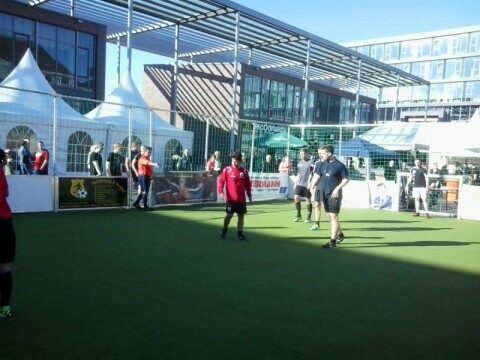 Mitarbeiter spielen Fußball auf einem mobilen Soccer Court bei einem Outdoor-Betriebsfest vor einem Firmengebäude