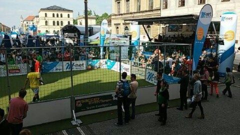 Stationärer Soccer Court mit weißen Banden, umlaufendem Netz und Überdachung, grüner Kunstrasen, Fußballtor mit Vereinsbranding in der Mitte, Innenansicht von der Längsseite